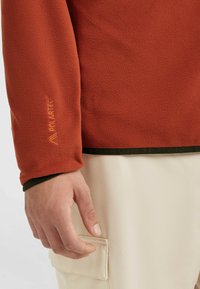 Roter Fleece-Pullover mit strukturierter Oberfläche, ausgestattet mit einem dunkelgrünen Saum und einem gestickten "Polartec"-Logo am Ärmel.