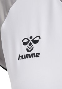 Hvid træningsskjorte med et sort "humme" logo og grafisk design. Stofet har en glat, struktureret overflade med grå detaljer.