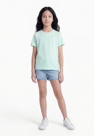 Dievča stojace, oblečené v svetlozelenom tričku s krátkym rukávom, rifľových šortkách a bielych teniskách, s dlhými tmavými vlasmi na jednoduchom bielom pozadí.