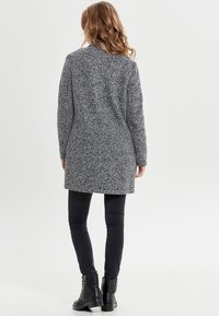 Kvinna med vågigt hår som bär en grå texturerad kappa, svarta tajta byxor och svarta ankelskotskor, vänd bort på en enfärgad vit bakgrund.