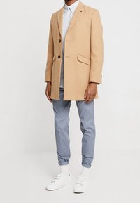 Mężczyzna w beżowym płaszczu, jasnoniebieskiej koszuli z guzikami, szarych spodni tapered, białych skarpetkach i białych trampkach stoi na białym tle.