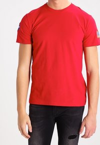 Hombre que lleva una camiseta roja de manga corta lisa con parches de la NASA y la bandera de EE. UU., combinada con jeans negros desgastados.