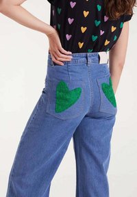 Persona que lleva vaqueros azules con parches de corazones verdes en los bolsillos traseros y una camiseta negra con estampados de corazones multicolores.