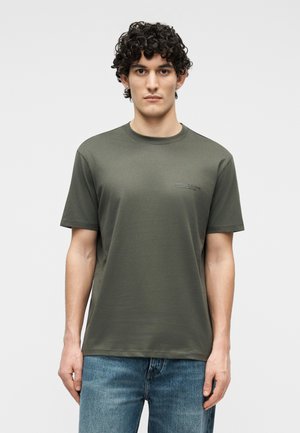 Άνδρας με σγουρά σκούρα μαλλιά, φορώντας χακί κοντομάνικο μπλουζάκι και μπλε τζιν, στέκεται μπροστά σε απλό λευκό φόντο.