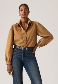 Brown corduroy button-up shirt with a collar, puffed sleeves, and front pocket, paired with high-waisted blue denim jeans and a black belt.