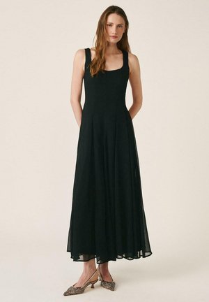 Mujer con vestido negro sin mangas hasta los tobillos y zapatos de punta con tacones pequeños, sobre un fondo claro y liso.