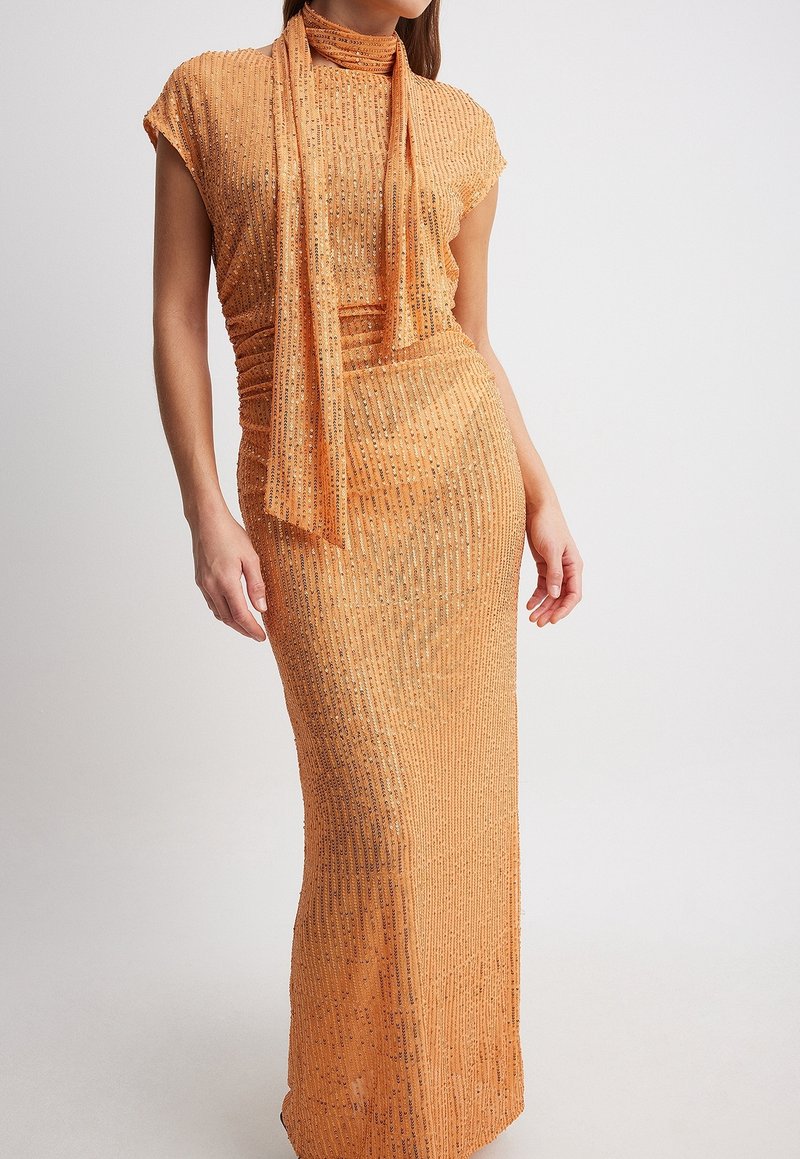 Femme portant une robe sans manches longue jusqu'au sol, en sequins pêche, avec une écharpe assortie drapée autour du cou, sur un fond uni.
