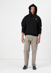 Schwarzer Hoodie mit Kordelzug und Logo, kombiniert mit hellgrauen Hosen. Das Model trägt marineblaue Schuhe und steht vor einem weißen Hintergrund.