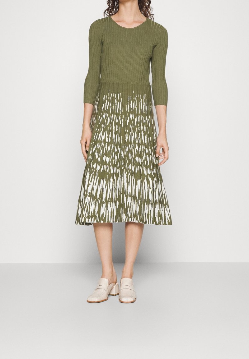 Robe en tricot avec manches trois-quarts, haut vert olive et ourlet blanc à motifs. Portée avec des chaussures beiges sans lacets. Texture lisse.
