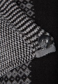 Stickad textil med ett svart och grått hundtandsmönster, med en rund fyra-håls knap i grå plast vid kanten.