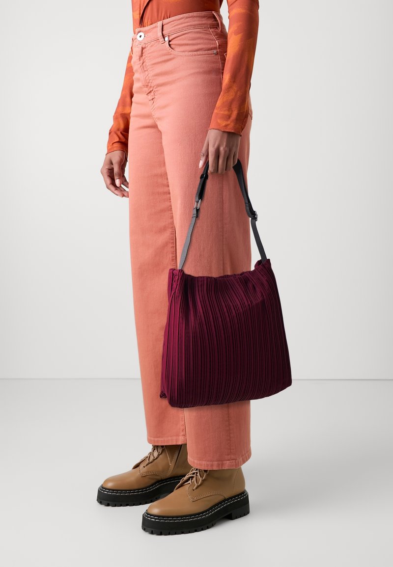 Sac à main plissé bourgogne avec un design structuré, des anses noires, associé à un pantalon évasé rose clair et des bottes à lacets brown.