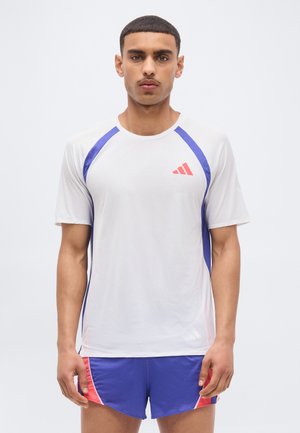 Mand stående med fronten mod kamera iført en hvid sportstrøje med blå accenter og blå shorts med røde detaljer mod en ensfarvet baggrund.