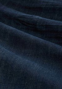 Tela de denim azul oscuro con textura visible y suaves pliegues que crean sombras y reflejos sutiles a lo largo de la superficie.
