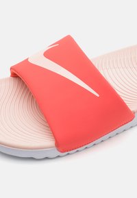 Rød slip-on sandal med en bred rem som har et hvitt Nike-logo, teksturert fotseng og en lys grå såle med bølgemønster.
