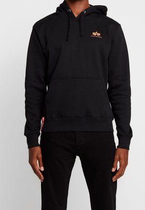 Mężczyzna noszący czarną bluzę z kapturem Alpha Industries z przednią kieszenią i logo na piersi, w połączeniu z czarnymi spodniami na jednolitym tle.