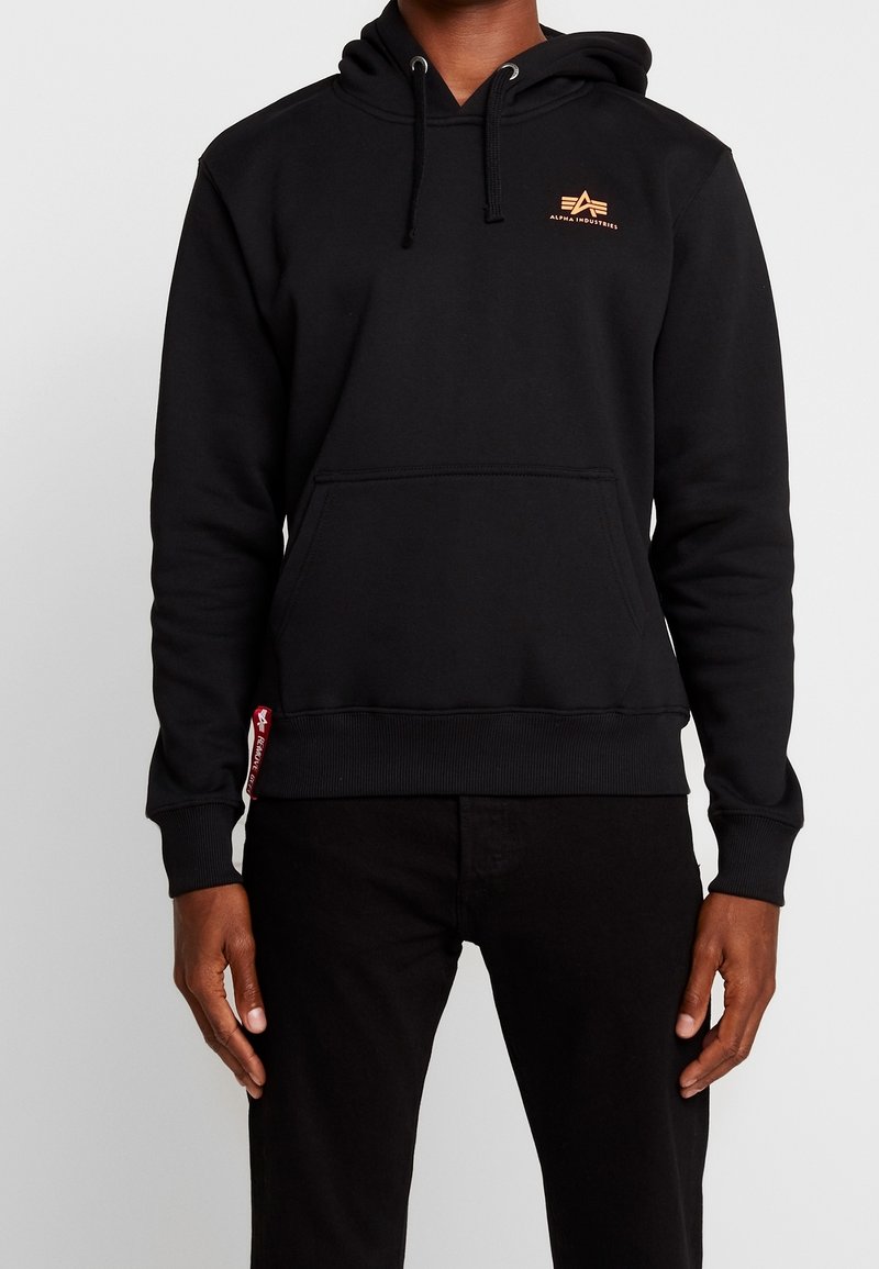 Homme portant un sweat à capuche noir Alpha Industries avec poche avant et logo sur la poitrine, associé à un pantalon noir sur un fond uni.