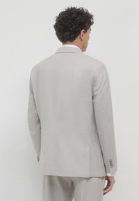 Ein Mann mit lockigem Haar, der einen hellbeigen Anzug trägt und mit dem Rücken zu einer schlichten weißen Wand steht.