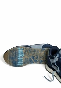 Marineblauwe en grijze suède sneaker met een zichtbare, geprofileerde zool waarop 'stuartgrift' staat, ligt op een witte achtergrond.