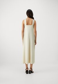 Pikk, ilma varrukat dress, helekreemikollane, ruudukujulise kaelusega ja voolava siluetiga, koos mustade plokk-kandadega sandaalidega.