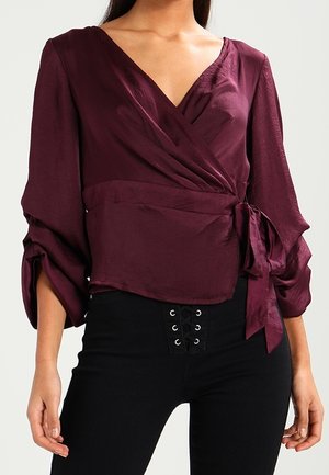 Frau trägt eine tiefviolette Wickelbluse mit langen Ärmeln und seitlichem Bindeband, kombiniert mit hoch taillierten schwarzen Hosen mit Schnür-Detail.