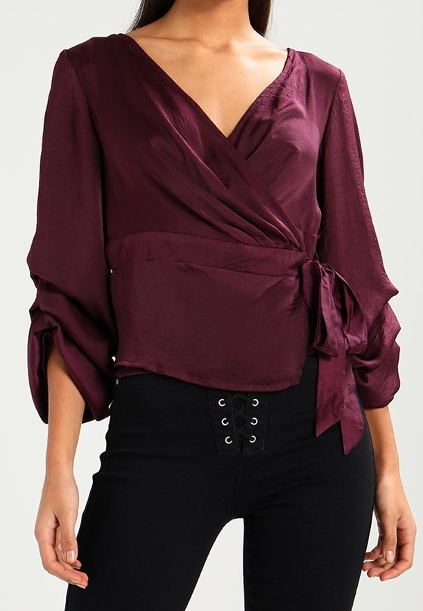 Femme portant un chemisier portefeuille violet profond à manches longues avec un lien sur le côté, associé à un pantalon noir taille haute avec un détail à laçage.