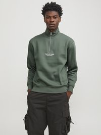 Sudadera verde con cremallera y cuello alto, que presenta un logo blanco en el pecho. Combinada con pantalones cargo negros, que muestran un ajuste relajado.