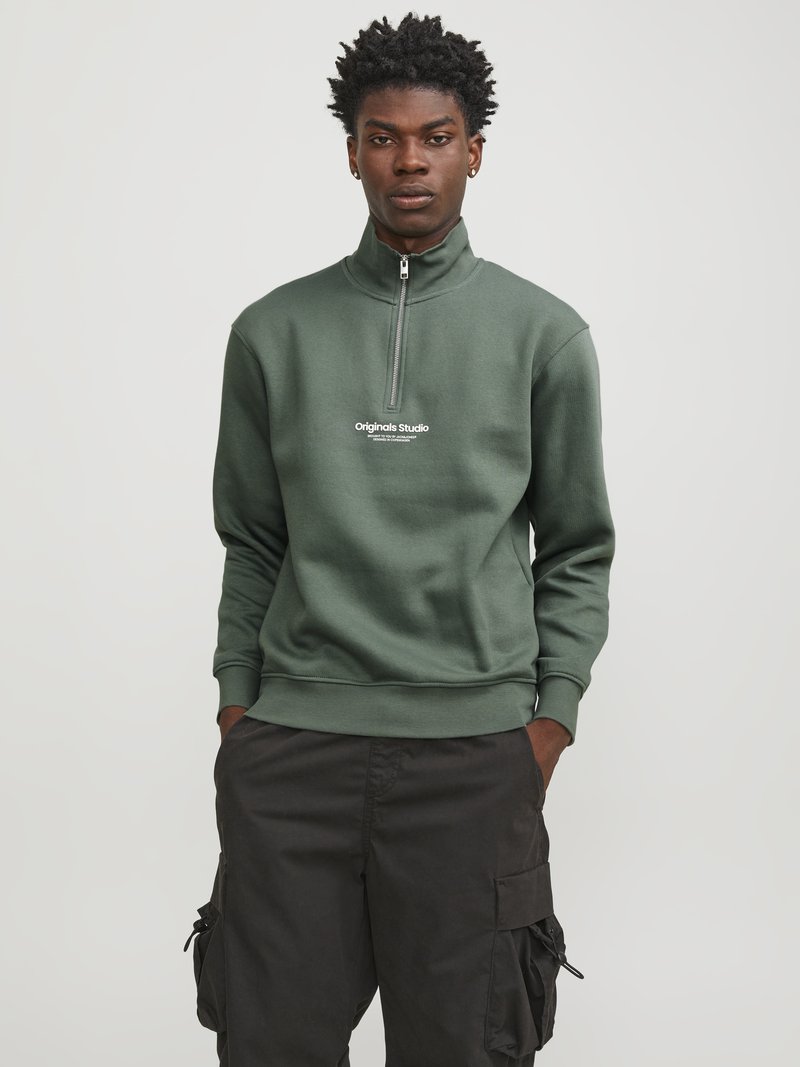Sudadera verde con cremallera y cuello alto, que presenta un logo blanco en el pecho. Combinada con pantalones cargo negros, que muestran un ajuste relajado.