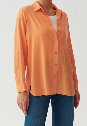 Femme portant une chemise orange à manches longues, ample et boutonnée, par-dessus un haut blanc et un jean bleu, debout devant un fond uni.