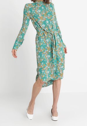 Teal floral midi dress with long sleeves, ruffled high neckline, side slit, fabric belt tied at waist, paired with white heeled shoes.