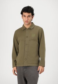 Olive green jacket made of sturdy fabric, featuring a button-up front, pointed collar, one chest pocket, and embroidered logo detail.