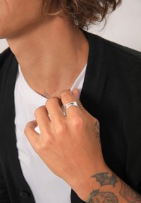 Silverring med lagerdesign bärs på en hand; huden visar tatueringar. Svart cardigan över en vit T-shirt; strukturerad halsringning synlig.