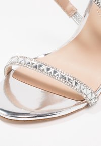 Sandale argentée à bout ouvert avec bride ornée de strass et semelle intérieure beige, présentée en gros plan.