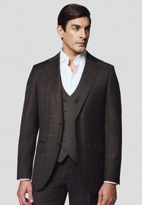 Brązowy garnitur w kratę z dopasowaną kamizelką, o kroju dopasowanym, z klapami w szpic. Biała koszula pod spodem; gładka faktura, czyste linie i trzy guziki.