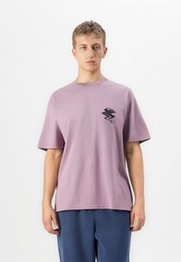 Noor allahõude juustega noormees kannab laia lavendlitootjaga T-särki, mille rinnal on väike must abstraktne muster, ja siniseid pükse.