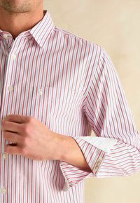 Biela košeľa na gombíky s červenými a sivými vertikálnymi pruhmi, obsahujúca vrecko na hrudi a kontrastnou bielou podšívkou na manžetách s etiketou.