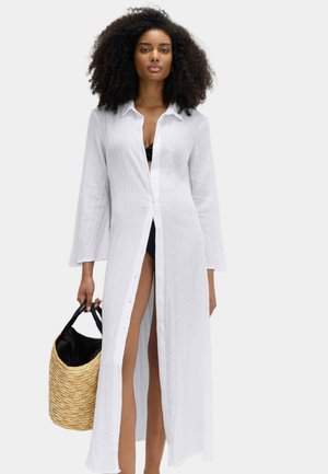 Kobieta o kręconych włosach, ubrana w długą białą sukienkę zapinaną na guziki, noszącą czarny strój kąpielowy, trzymająca plecioną słomianą torbę z czarnymi rączkami.