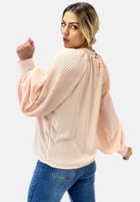 Blusa rosa claro con mangas plisadas, textura suave, cuello redondo y pequeño cierre de botón en la parte posterior. Llevada con jeans azules.