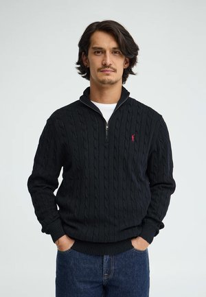 Uomo con capelli scuri di media lunghezza, indossa un maglione nero a trecce con zip a un quarto e jeans blu, con le mani in tasca e un'espressione neutra.
