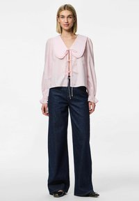 Blusa rosa chiaro con un ampio colletto arruffato, legato davanti con un nastro abbinato, abbinata a pantaloni di jeans scuri a gamba larga.