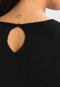 Haut en tricot noir avec une petite ouverture en forme de clé dans le dos, fermée par un bouton noir, révélant un tatouage en forme de cœur sur la peau.
