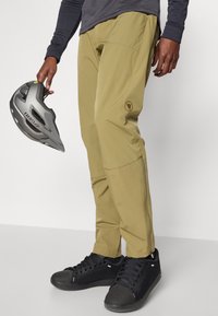 Khaki vonkajšie nohavice vyrobené z ľahkého materiálu, s vreckami na zips a logom. V ruke sú čierne športové topánky a šedá prilba.
