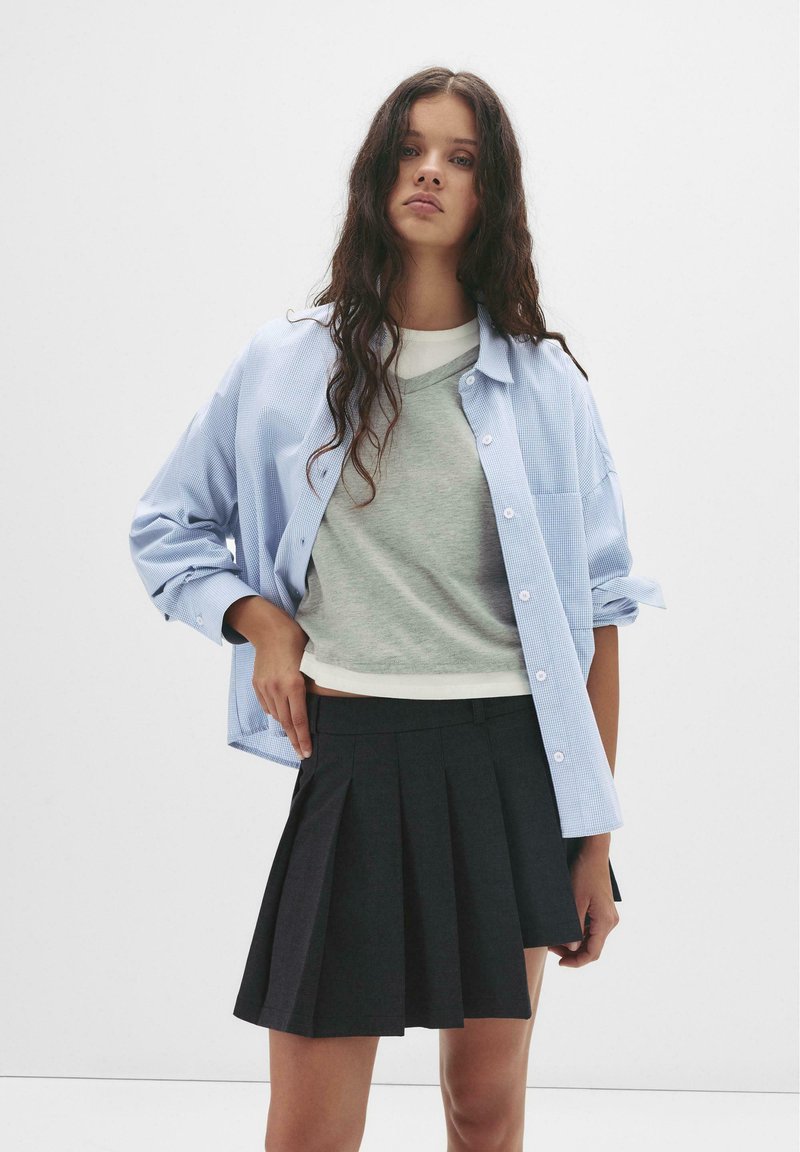 Jasnoniebieska koszula w kratę na szaro-swetrowym V-neck, zestawiona z czarną plisowaną mini spódnicą. Gładki materiał, dopasowany krój, prosty design.