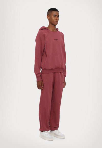 Conjunto de moletom com capuz e calças de treino na cor bordeaux, feito de material suave, com um corte descontraído e um pequeno logo impresso no peito. Usado com sapatilhas brancas.