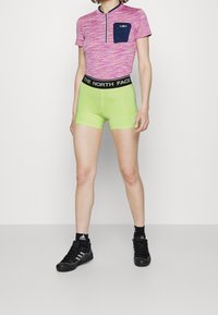 T-shirt de sport à manches courtes, multicolore, avec une fermeture éclair et une poche bleu marine, associé à un short vert lime avec une taille noire. Chaussures de sport noires.