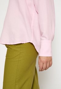 Persona che indossa una camicia lunga rosa larga e pantaloni verde oliva, con il braccio destro rilassato al suo fianco su uno sfondo neutro.