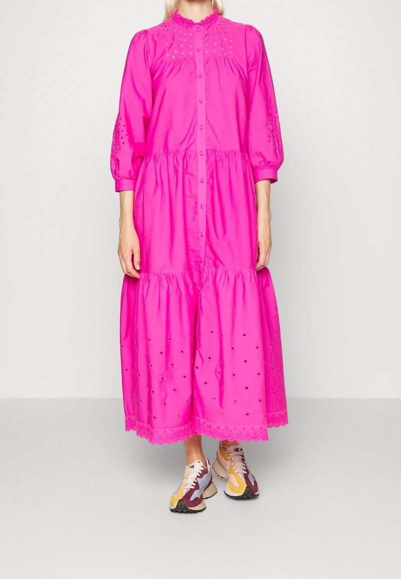 Robe longue en coton rose avec manches bouffantes, boutonnage avant, col à volants et détails en broderie anglaise. Associée à des chaussures de sport colorées.