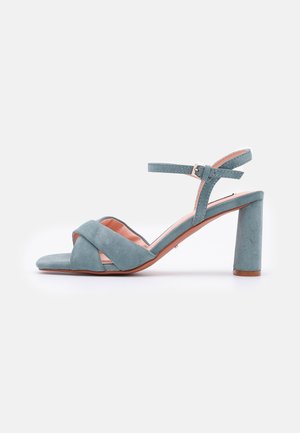Sandały na wysokim obcasie w jasnoniebieskim zamszu z kwadratowym noskiem i szerokim, blokowym obcasem. Posiadają regulowany pasek w okolicy kostki oraz krzyżujący się przód.