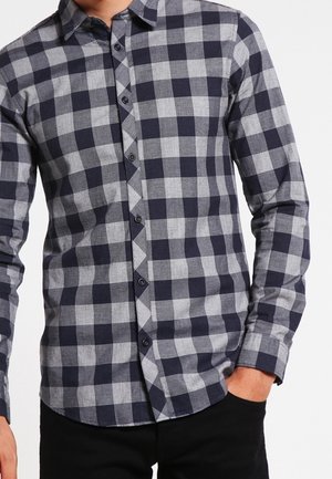 Homme portant une chemise ajustée à manches longues boutonnée avec un motif à carreaux bleu marine et gris, main partiellement dans la poche d'un pantalon noir.