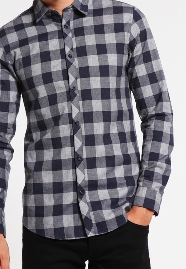 Homme portant une chemise ajustée à manches longues boutonnée avec un motif à carreaux bleu marine et gris, main partiellement dans la poche d'un pantalon noir.