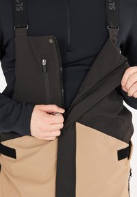 Bavaglini impermeabili neri e panna con tasche anteriori con cerniera e spallacci regolabili, realizzati in un tessuto testurizzato e con dettagli in velcro.